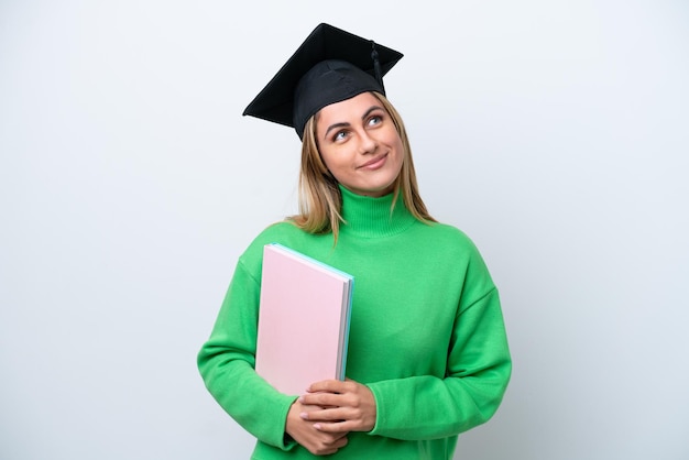 Junge Frau mit Universitätsabsolvent isoliert auf weißem Hintergrund, die eine Idee denkt, während sie nach oben schaut