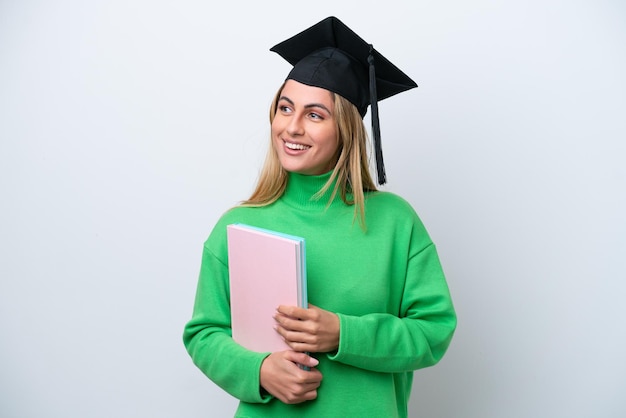 Junge Frau mit Universitätsabsolvent isoliert auf weißem Hintergrund, die zur Seite schaut und lächelt