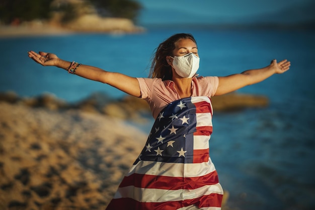 Junge Frau mit US-Nationalflagge und schützender N95-Maske am Strand, während sie die Ausbreitung der Epidemie verhindert und Coronavirus und Pandemie COVID-19 behandelt.