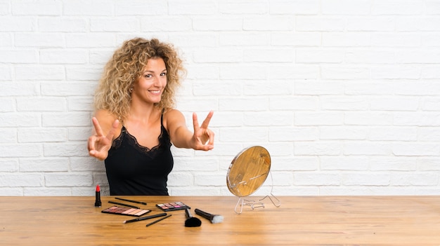 Junge Frau mit vielen Make-upbürste in einer Tabelle lächelnd und Siegeszeichen zeigend