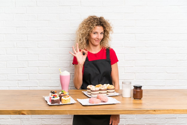 Junge Frau mit vielen verschiedenen Minikuchen in einer Tabelle, die ein okayzeichen mit den Fingern zeigt