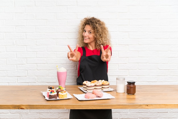 Junge Frau mit vielen verschiedenen Minikuchen in einer Tabelle lächelnd und Siegeszeichen zeigend