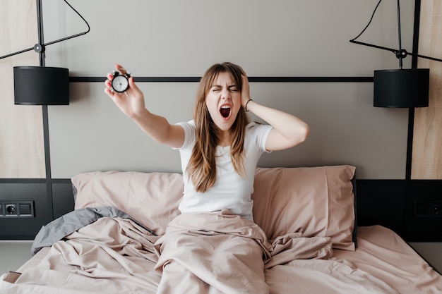 Junge Frau mit Wecker in der Hand, die am Morgen im Bett zu Hause aufwacht