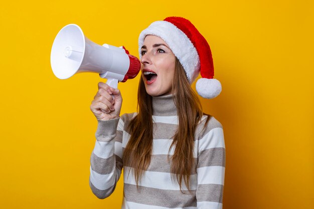 Junge Frau mit Weihnachtsmannmütze spricht in ein Megaphon auf gelbem Hintergrund