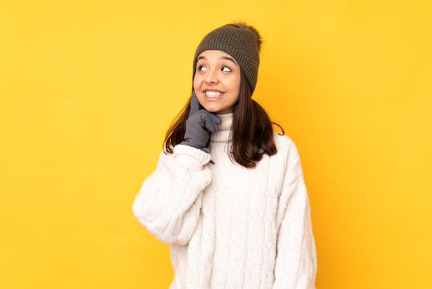 Junge Frau mit Winterhut über gelber Wand eine Idee beim oben schauen denkend