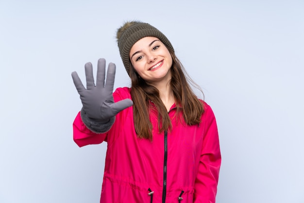 Junge Frau mit Wintermütze lokalisiert auf blauer Wand, die fünf mit den Fingern zählt