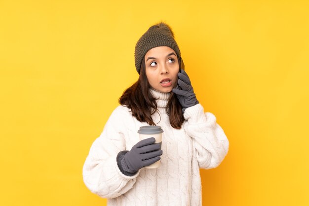 Junge Frau mit Wintermütze über isolierter gelber Wand, die Kaffee hält, um und ein Handy wegzunehmen