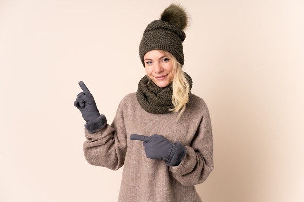 Junge Frau mit Wintermütze über isolierter Wand, die Finger zur Seite zeigt