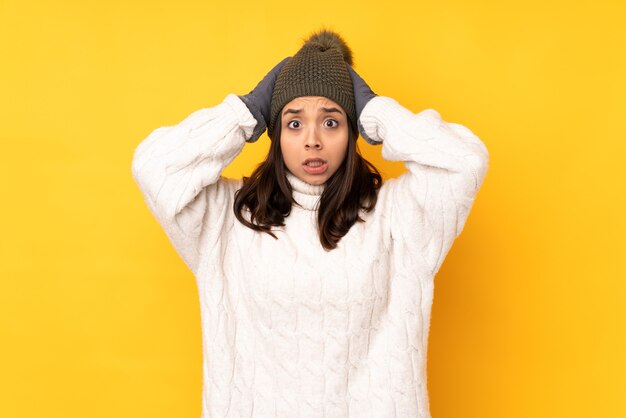 Junge Frau mit Wintermütze über lokalisiertem gelbem Hintergrund frustriert und nimmt Hände auf Kopf