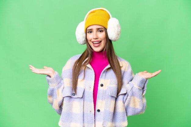 Junge Frau mit Wintermuffs vor isoliertem Chroma-Key-Hintergrund mit schockiertem Gesichtsausdruck