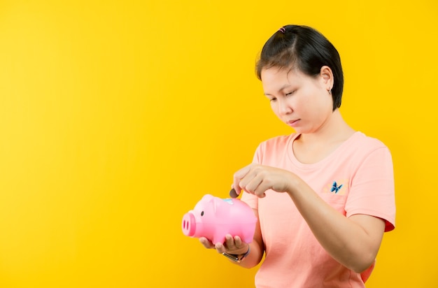 Junge Frau münzbetrieben glücklich mit rosa Sparschwein, einem gelben Hintergrund. Konzepteinsparung.