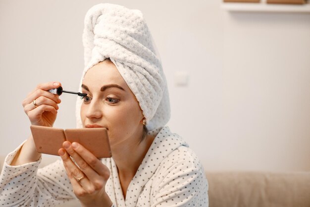 Junge Frau nach dem Duschen mit weißem Morgenmantel hat ein weißes Handtuch, das ihr Haar umwickelt Mädchen sitzt auf dem Sofa und macht sich vor dem Spiegel im hellen Innenraum Frau, die eine Mascara anwendet