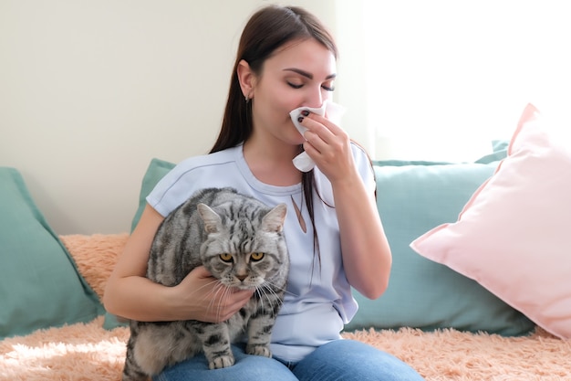 Junge Frau niest von Pelzallergie auf dem Sofa und spielt mit ihrer Katze.