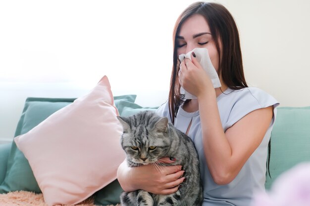 Junge Frau niest von Pelzallergie auf dem Sofa und spielt mit ihrer Katze.