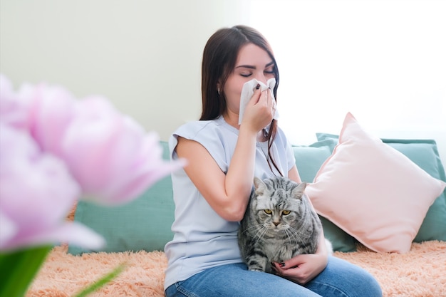 Junge Frau niest von Pelzallergie auf dem Sofa und spielt mit ihrer Katze.