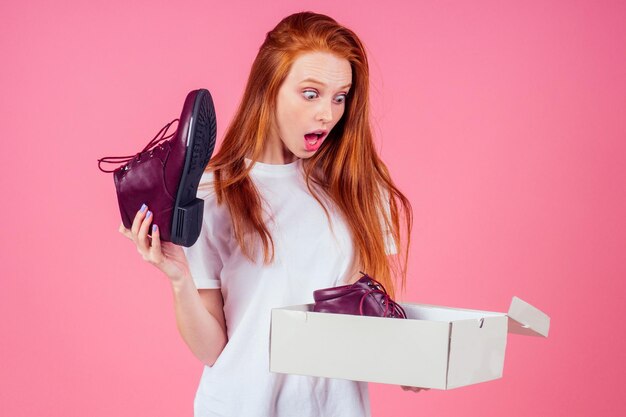 Foto junge frau öffnet paket im studio rosa hintergrundherbst winter rote lederstiefel saleshe trägt weißes baumwollhemd