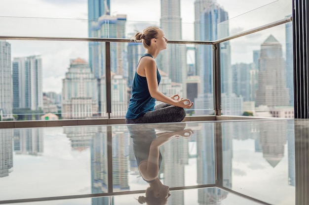 Junge Frau praktiziert morgens Yoga auf ihrem Balkon mit Panoramablick auf die Stadt und die Wolkenkratzer