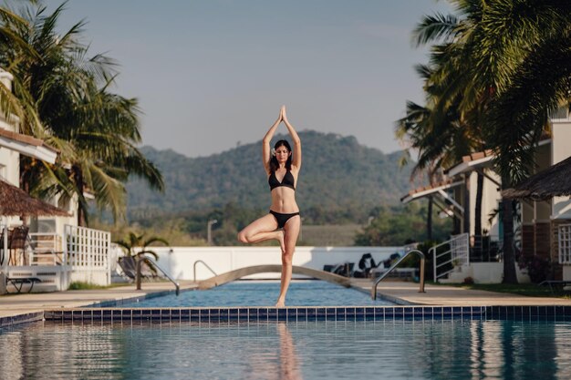 Junge Frau praktiziert Posen-Yoga am Swimmingpool und am Naturstrand bei Sonnenuntergang oder Sonnenaufgang Yoga und gesundes Konzept Yoga in Baumhaltung