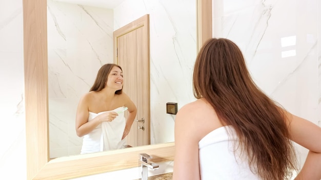 Junge Frau putzt die Zähne mit einer weißen Zahnbürste und betrachtet die Spiegelung im Badezimmerspiegel mit einem Lächeln aus der Nähe