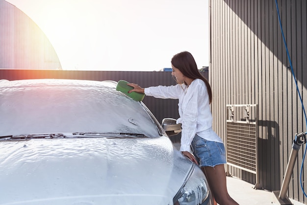 Junge Frau reinigt ihr Auto mit Schaum mit einem Schwamm in der Autowasche