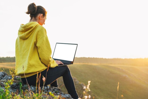 Junge Frau, Reisende, Freiberuflerin in gelbem Hoodie mit geöffnetem Laptop mit weißem Bildschirm auf dem Hintergrund der schönen Aussicht