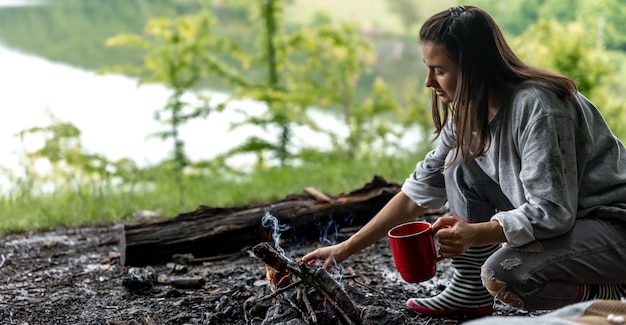 Junge Frau ruht in der Nähe des Feuers mit einer Tasse wärmenden Getränks im Wald in der Nähe des Flusses