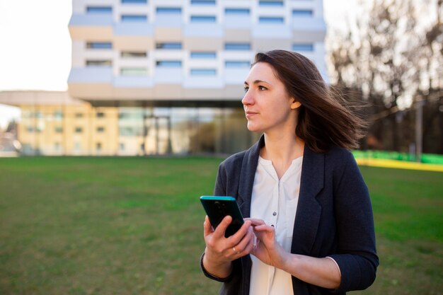 Junge Frau schaut auf das Telefon, telefoniert, spricht auf Video, lächelt, winkt, grüßt, auf der Straße im Park, in der Nähe des Geschäftszentrums