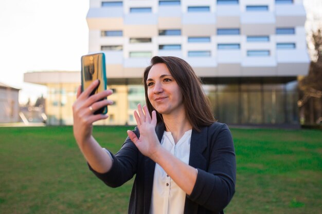 Junge Frau schaut auf das Telefon, telefoniert, spricht auf Video, lächelt, winkt, grüßt, auf der Straße im Park, in der Nähe des Geschäftszentrums