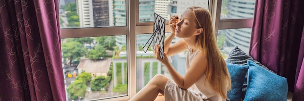 Junge Frau schminkt sich am Fenster mit Panoramablick auf die Wolkenkratzer und die Großstadt BANNER LANGFORMAT