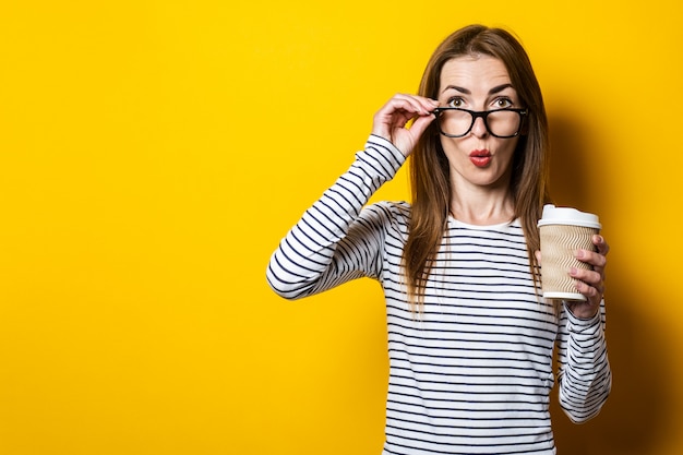 Junge Frau schockiert in Gläsern mit einer Pappbecher Kaffee auf gelbem Grund