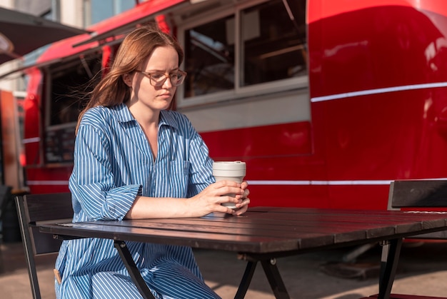 Junge Frau sitzt allein mit zusammenklappbarer Öko-Kaffeetasse in der Nähe von Street Food Truck City Lifestyle