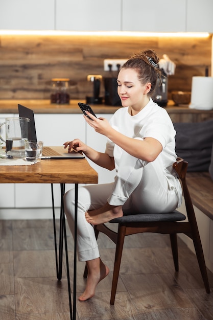 Junge Frau sitzt am Küchentisch mit einem Laptop und spricht auf einem Handy und lächelt. Erfolgreiches Mädchen, das zu Hause lacht und arbeitet. Schöne stilvolle Frau, die zu Hause lächelt und sich entspannt.