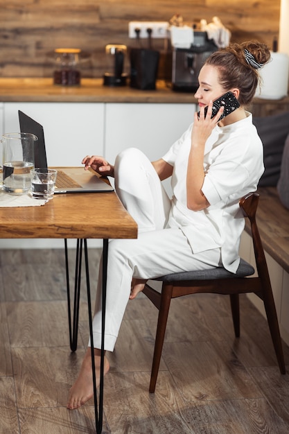 Junge Frau sitzt am Küchentisch mit einem Laptop und spricht auf einem Handy und lächelt. Erfolgreiches Mädchen, das zu Hause lacht und arbeitet. Schöne stilvolle Frau, die zu Hause lächelt und sich entspannt.