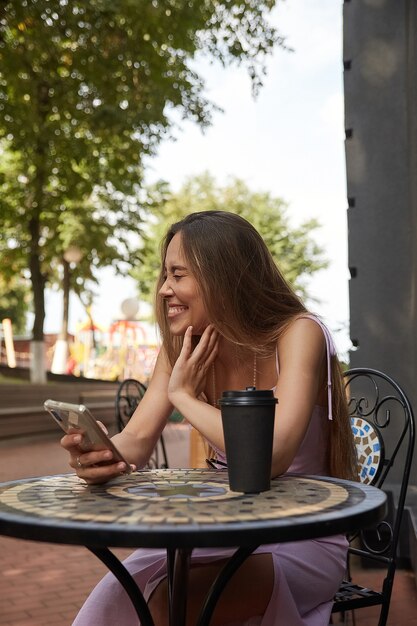 Junge Frau sitzt am Tisch mit Getränk, hält Smartphone und lacht. Dame, die eine Tasse Kaffee außerhalb des Cafés trinkt, im Internet auf dem Handy surft, online chattet. moderne Kommunikationstechnik