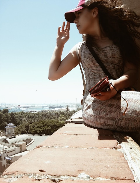 Junge Frau sitzt an einem sonnigen Tag an einer Stützmauer gegen den Himmel