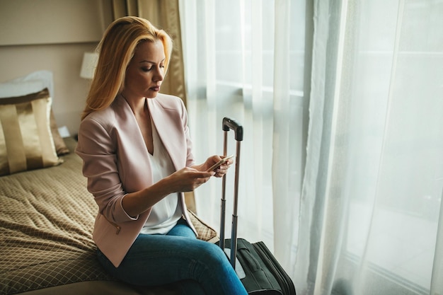 Junge Frau sitzt auf dem Bett in einem Hotelzimmer und hält ein Mobiltelefon in der Hand