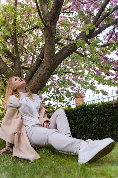Junge Frau sitzt auf dem Gras unter Kirschblüten und schaut auf Mädchen ruht im Frühlingspark unter blühendem Baum Vertikaler Rahmen