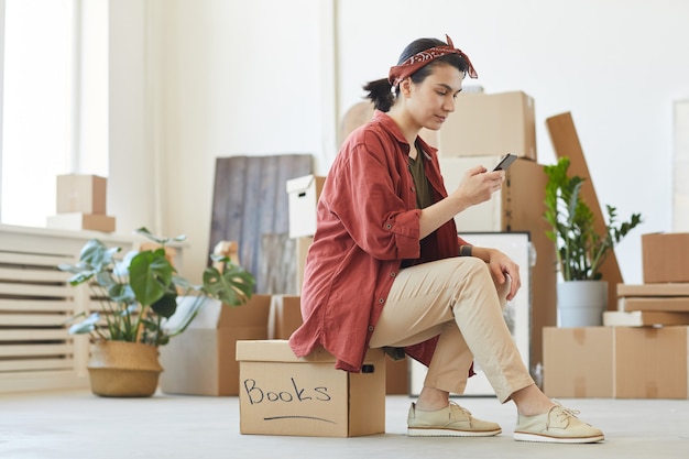 Junge Frau sitzt auf der Box und benutzt ihr Handy, um das Auto für den Transport von Dingen zu bestellen