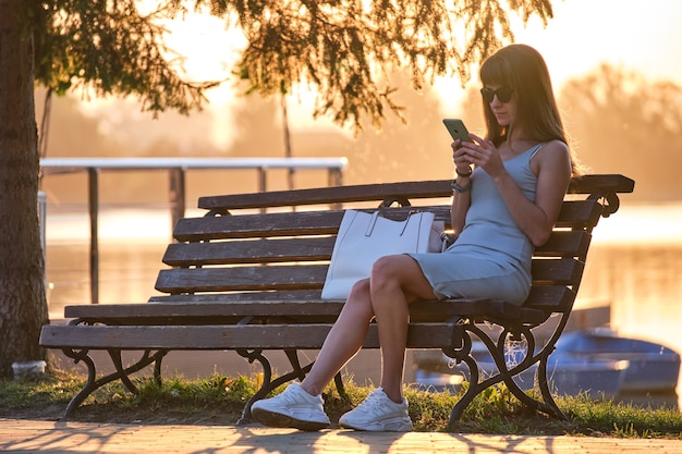 Junge Frau sitzt auf der Parkbank, die an einem warmen Sommerabend ihr Handy im Freien durchsucht. Kommunikations- und mobiles Verbindungskonzept.