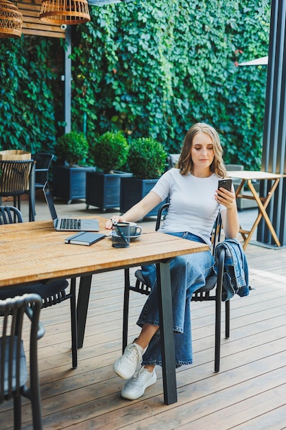 Junge Frau sitzt auf der Terrasse eines Cafés auf der Straße eines modernen Cafés. Frau sitzt am Tisch mit Telefon und benutzt Laptop im Freien. Fernarbeit in einem Sommercafé