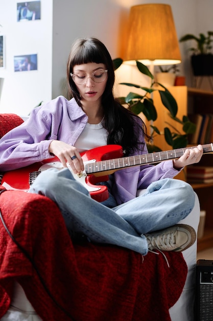 Junge Frau sitzt auf einem Sessel im Zimmer und spielt Gitarre