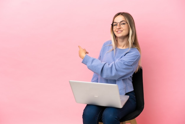 Junge Frau sitzt auf einem Stuhl mit Laptop über isoliertem rosa Hintergrund, der nach hinten zeigt