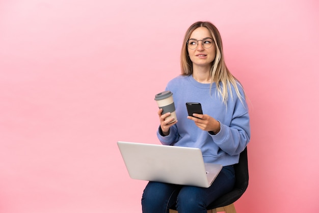 Junge Frau sitzt auf einem Stuhl mit Laptop über isoliertem rosa Hintergrund und hält Kaffee zum Mitnehmen und ein Handy, während sie etwas denkt