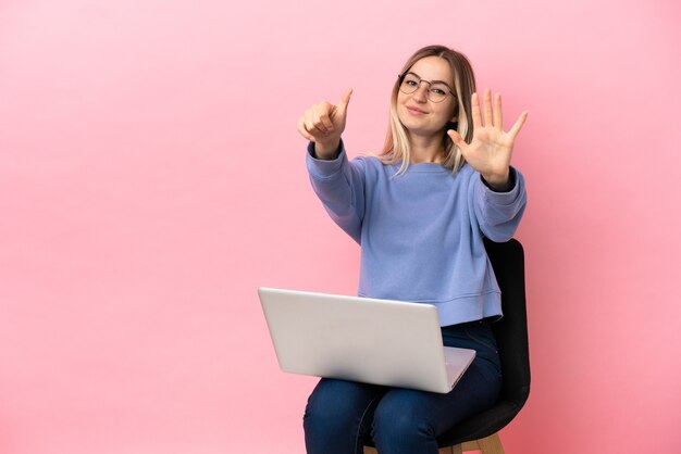 Junge Frau sitzt auf einem Stuhl mit Laptop über isoliertem rosa Hintergrund und zählt sechs mit den Fingern