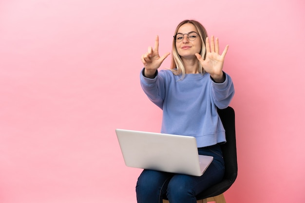 Junge Frau sitzt auf einem Stuhl mit Laptop über isoliertem rosa Hintergrund und zählt sieben mit den Fingern