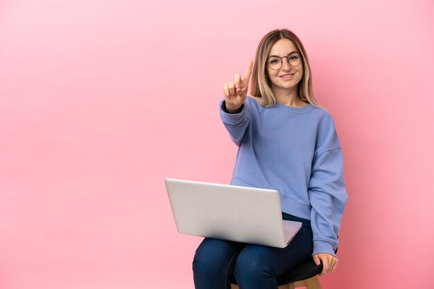 Junge Frau sitzt auf einem Stuhl mit Laptop über isoliertem rosa Hintergrund und zeigt und hebt einen Finger