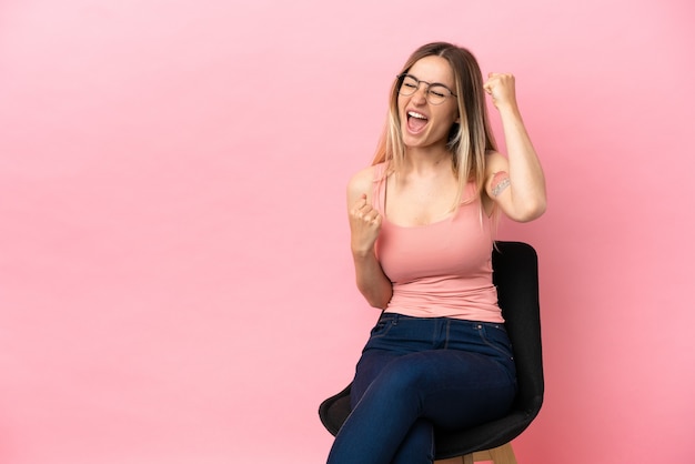 Junge Frau sitzt auf einem Stuhl über isoliertem rosa Hintergrund und feiert einen Sieg