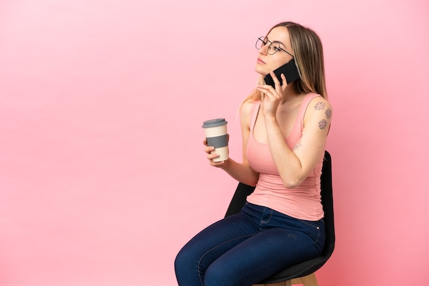 Junge Frau sitzt auf einem Stuhl über isoliertem rosa Hintergrund und hält Kaffee zum Mitnehmen und ein Handy