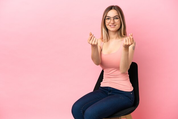 Junge Frau sitzt auf einem Stuhl über isoliertem rosa Hintergrund und macht Geldgeste