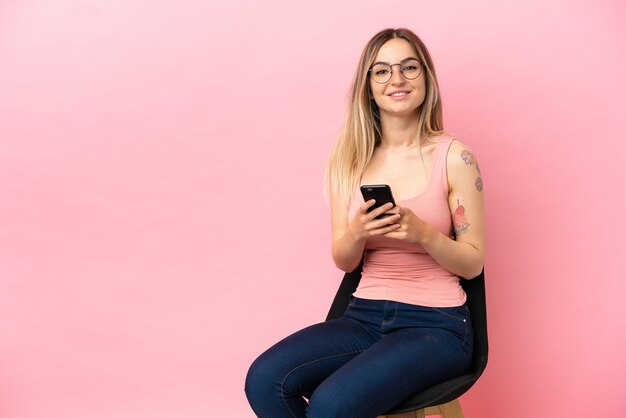 Junge Frau sitzt auf einem Stuhl über isoliertem rosa Hintergrund und sendet eine Nachricht mit dem Handy